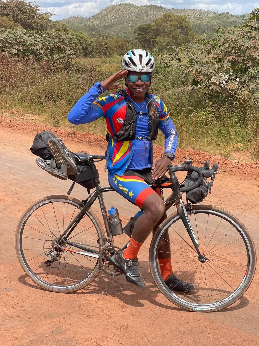 Miguel Masaisai in cycling clothes standing with his bicycle on a dirt road, saluting.